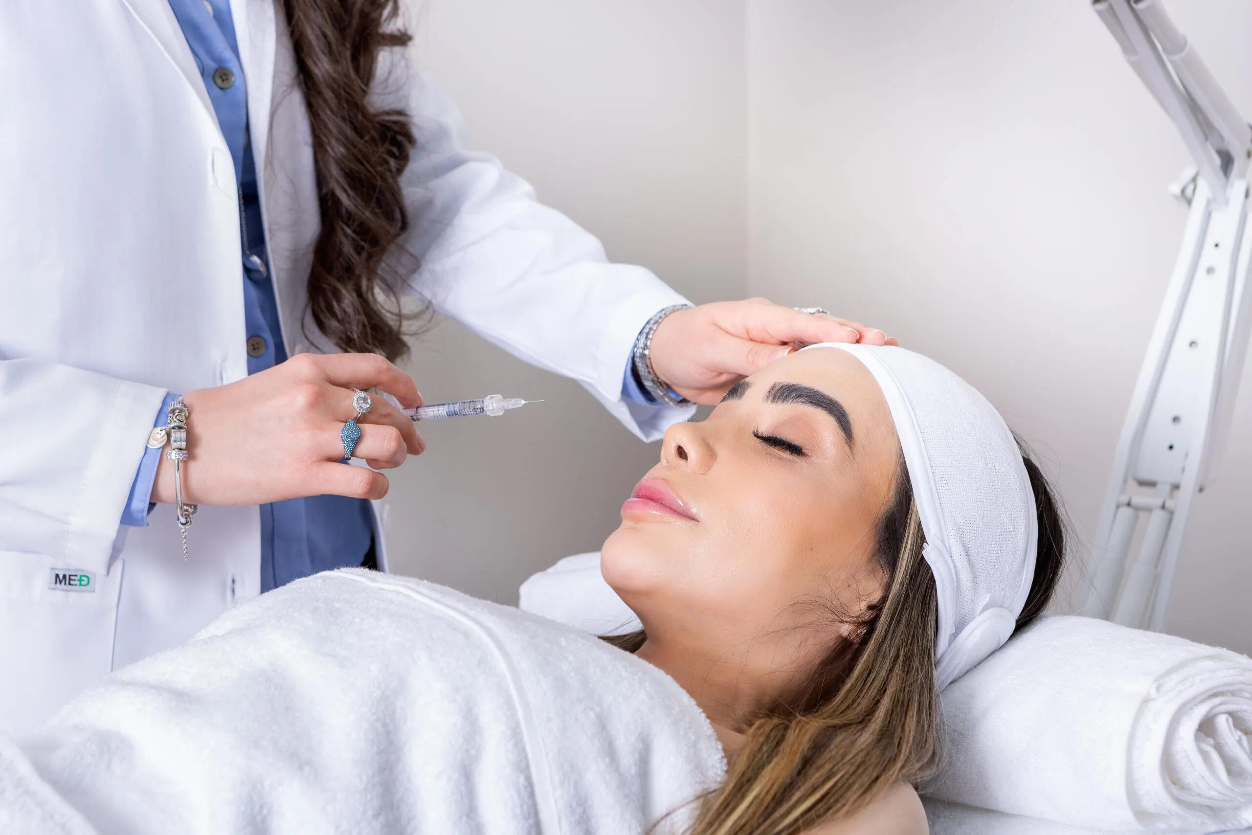 La doctora aplica un tratamiento inyectable en la frente de una paciente recostada.
